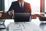 Businessperson in a suit points to a laptop and blueprints on a conference table during a planning meeting.